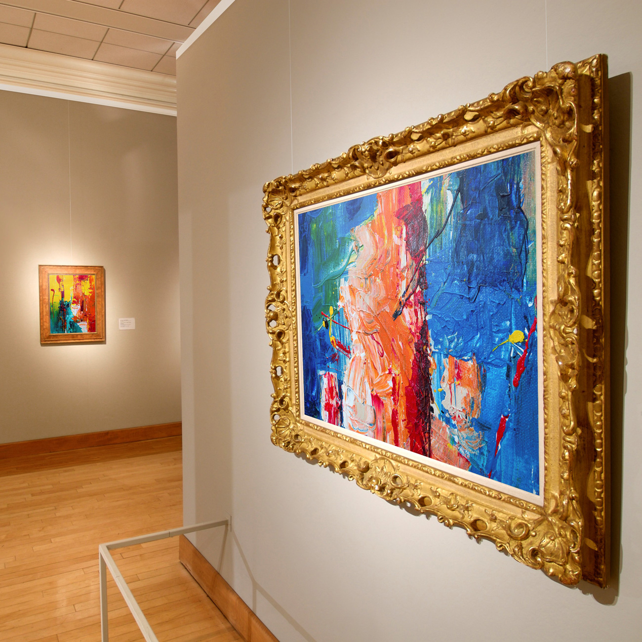 Large abstract painting in a gilded ornate frame hung on a gray gallery wall, with a smaller colorful piece in the background.