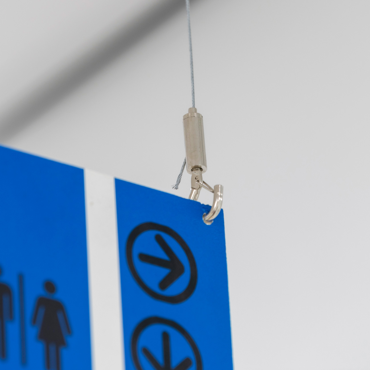 Close-up of a blue hanging sign attached by a metal clip to a gray wire, with restroom pictograms visible on the sign.