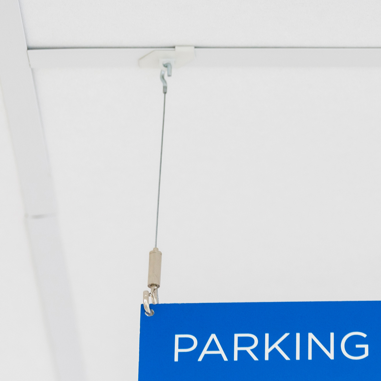Blue PARKING sign hanging from a thin cord attached to the ceiling, partially visible in the frame, inside a building.
