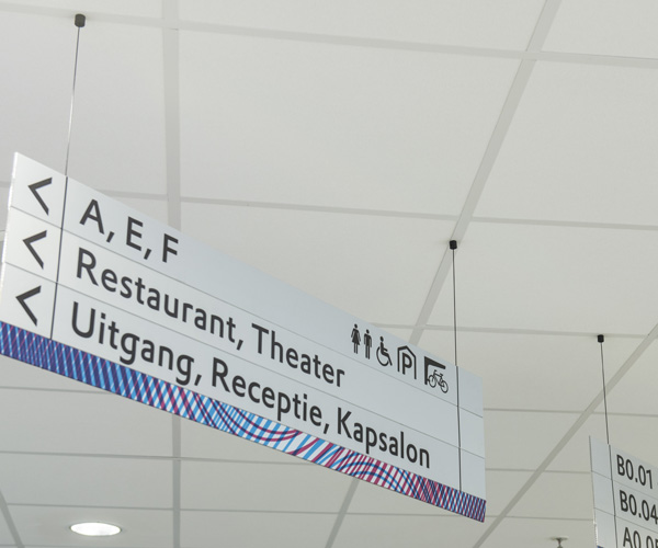 Hang­ing directional sign in a bright interior showing A, E, F; Restaurant, Theater; Uitgang, Receptie, Kapsalon with accessibility icons and a blue striped edge.